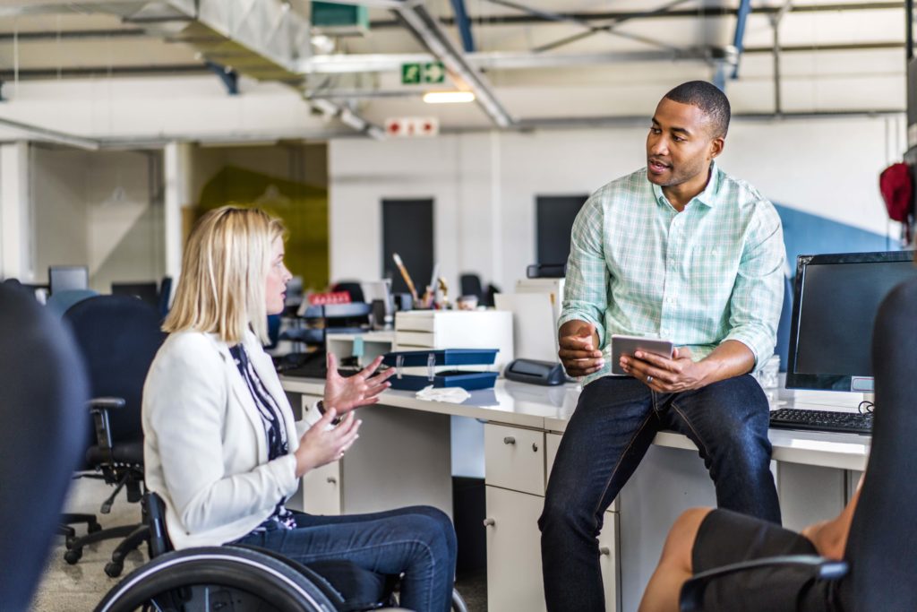 A photo of disabled businesswoman sharing ideas with coworker. Male professional is holding digital tablet. They are in creative office.